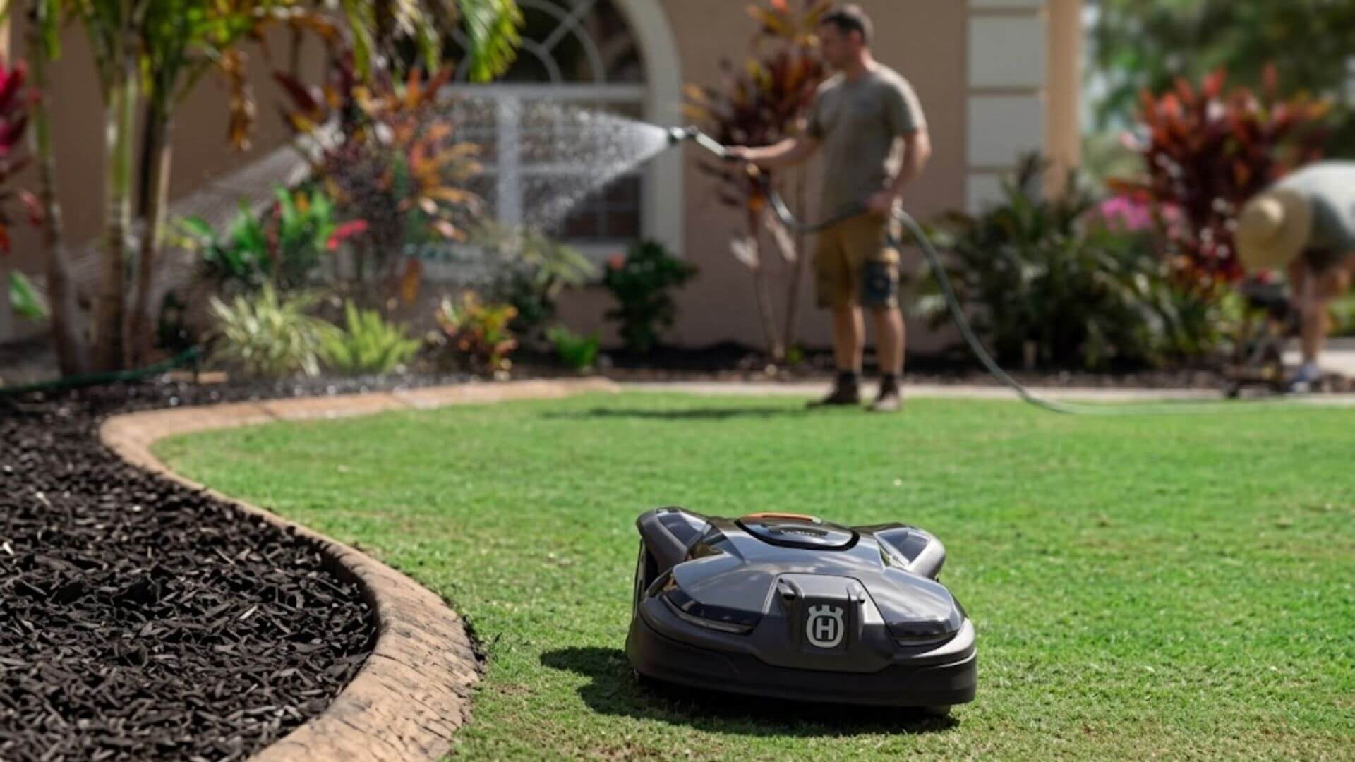 Husqvarna Automower mowing a residential lawn while a homeowner waters plants in the background.
