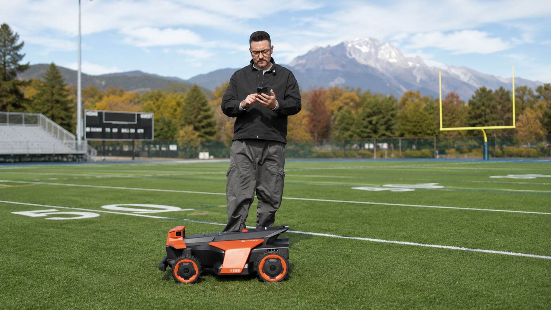 Grounds manager using a smartphone to monitor a Kress robotic mower maintaining an athletic field with mountain scenery in the distance.