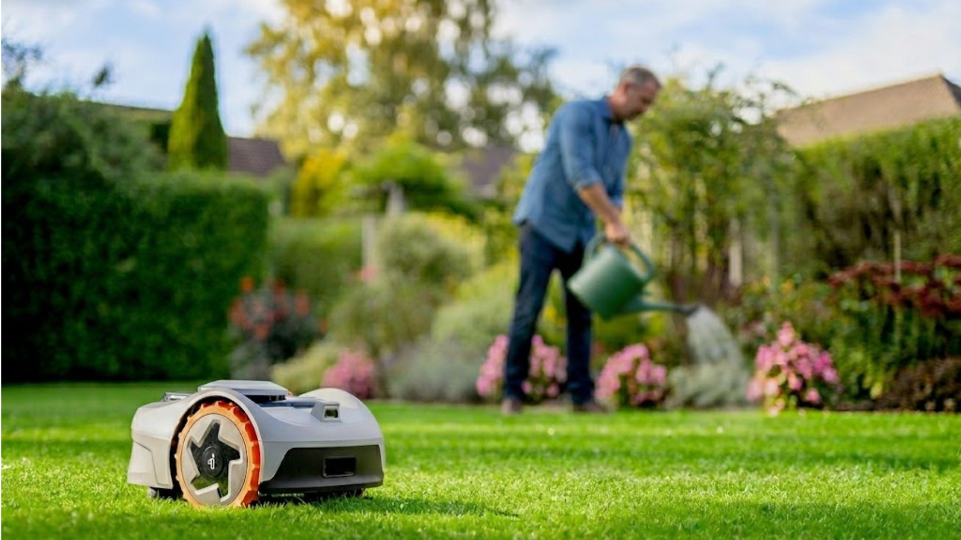 Navimow robotic mower operating on a residential lawn while a homeowner waters flower beds in the background.