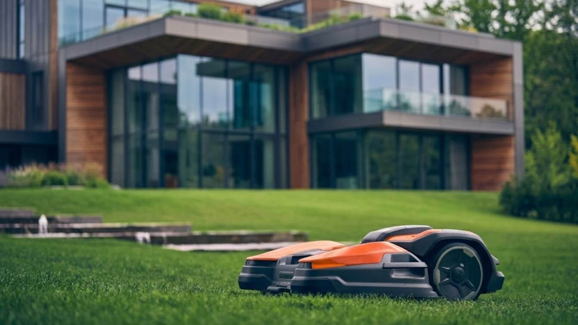 Husqvarna robotic mower mowing a manicured lawn in front of a contemporary luxury home with large glass windows.